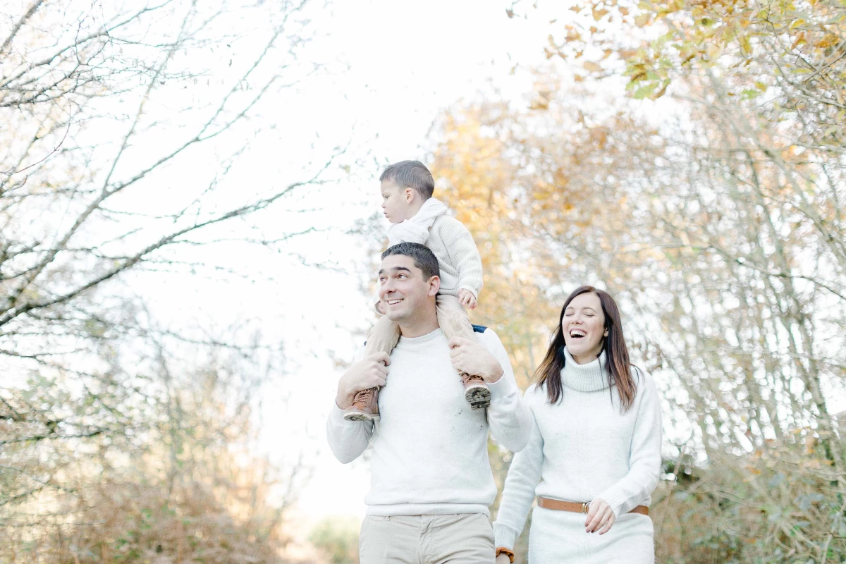 Séance famille en automne : père portant son fils sur les épaules, mère souriante à côté, vêtus de blanc, dans une allée d'arbres dorés, ambiance chaleureuse et complice