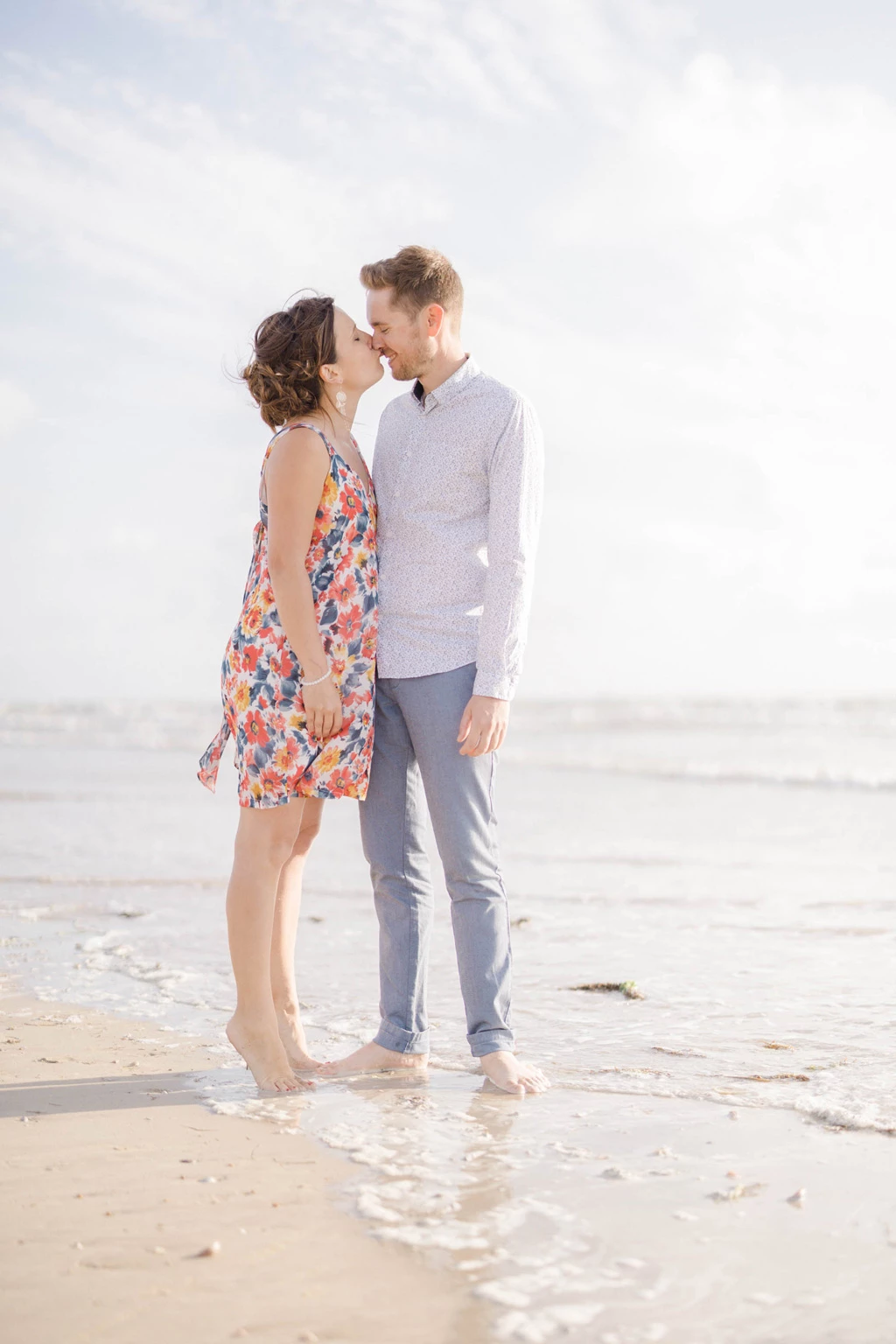 Couple s'embrassant pieds nus sur une plage, elle en robe fleurie, lui en chemise blanche