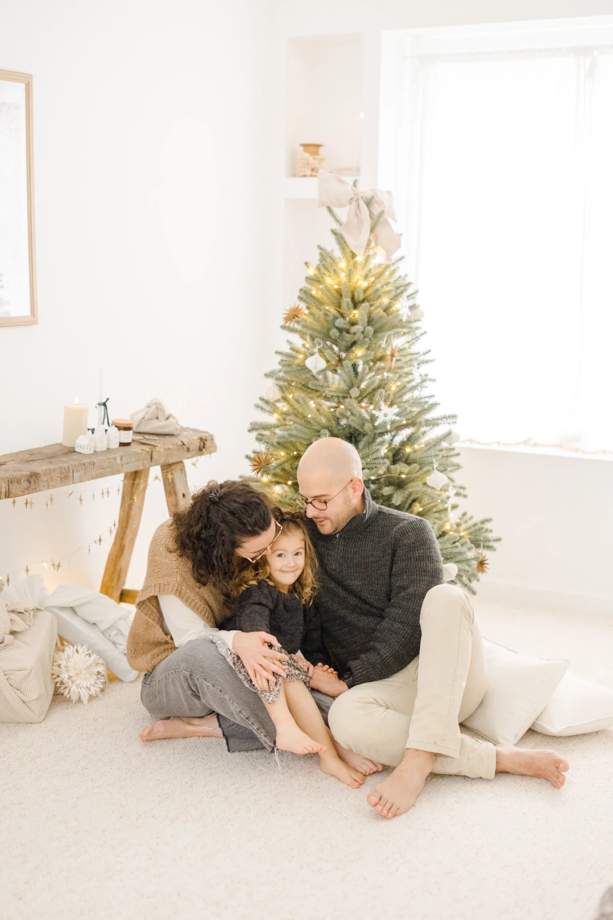 Parents enlacent tendrement leur fille devant un sapin de Noël illuminé dans un intérieur blanc épuré