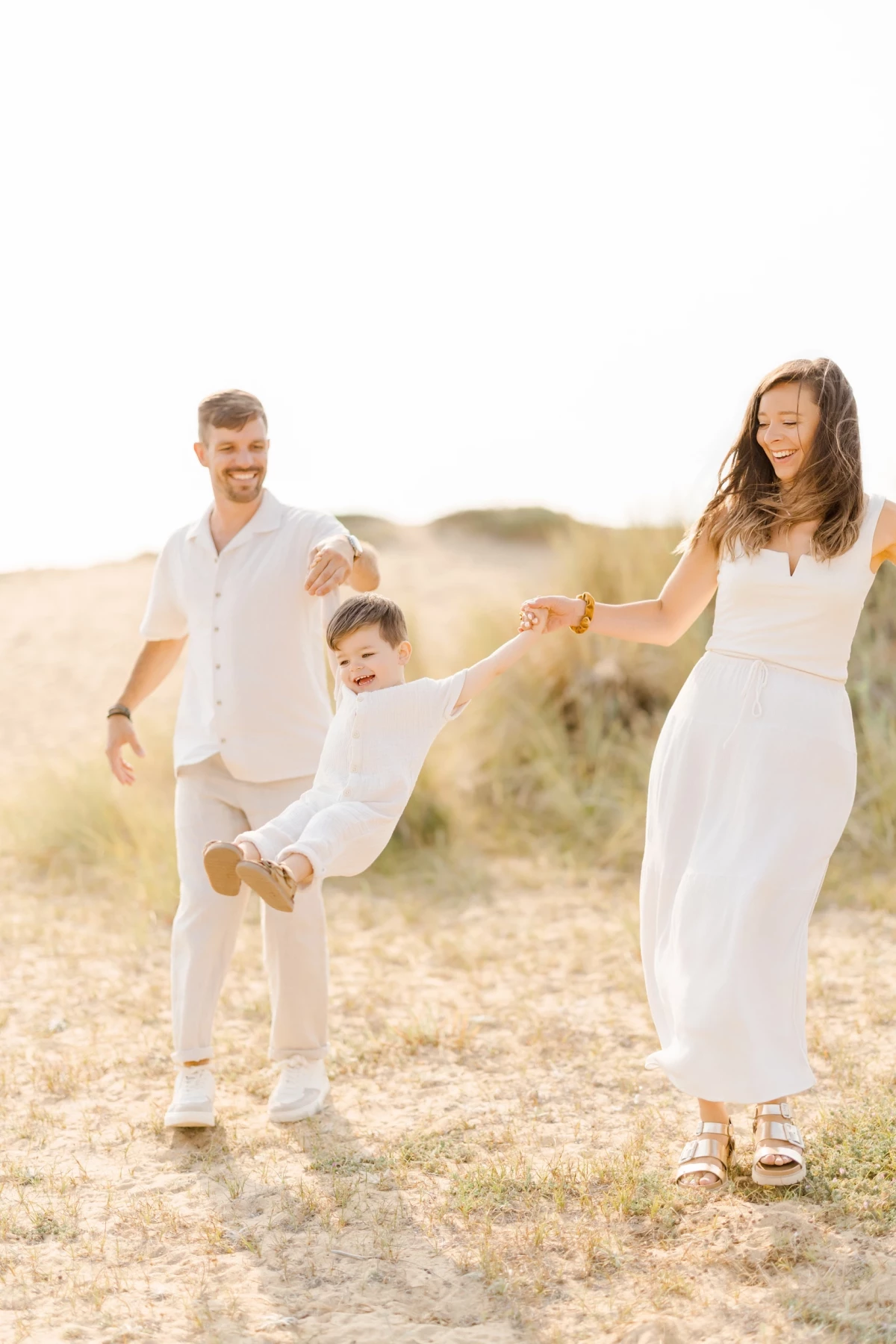 Séance famille sur la plage en Vendée : parents balancent leur enfant avec joie dans les dunes sous la lumière dorée du soleil