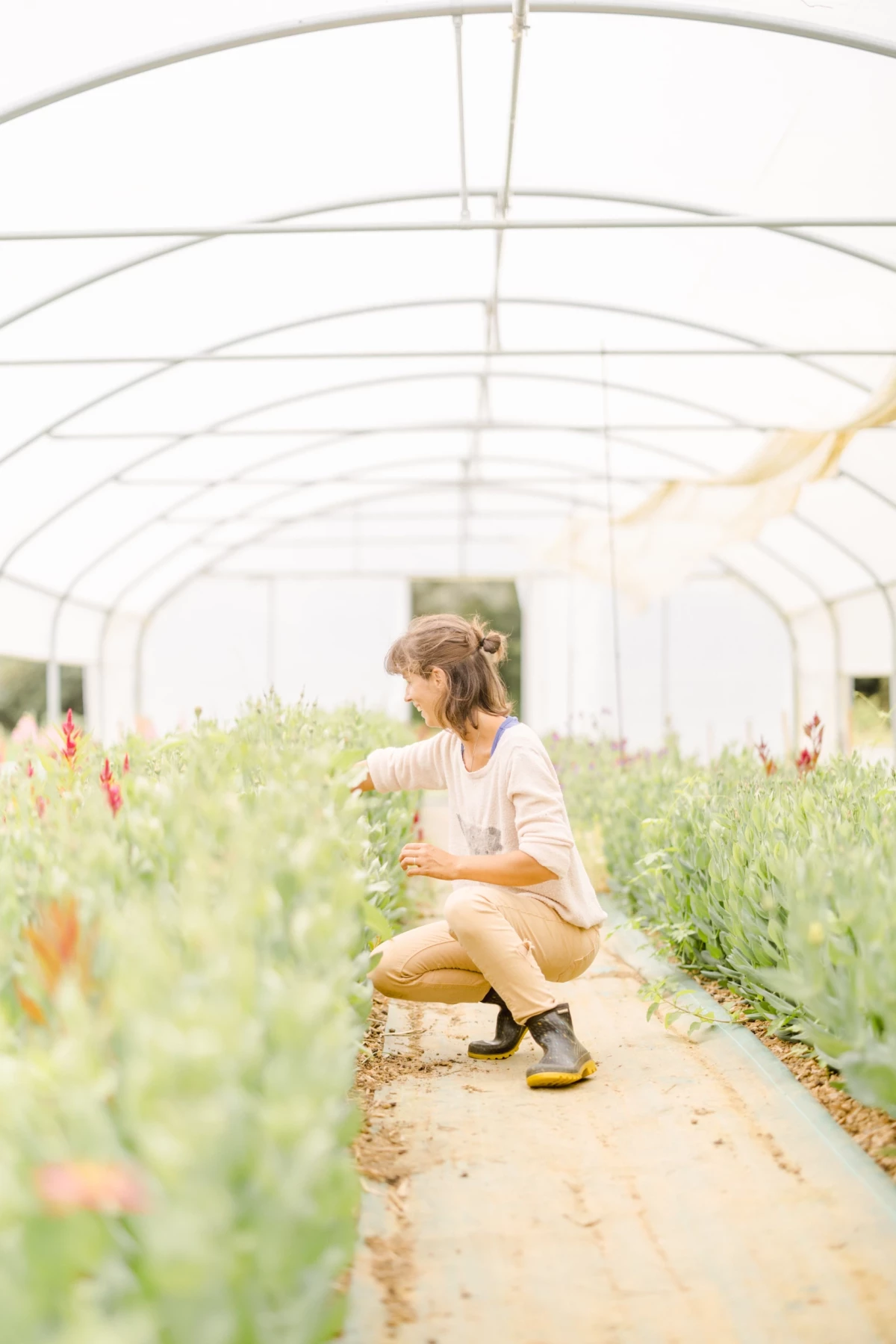 Femme en tenue beige accroupie dans une serre florale, inspectant des fleurs sauvages