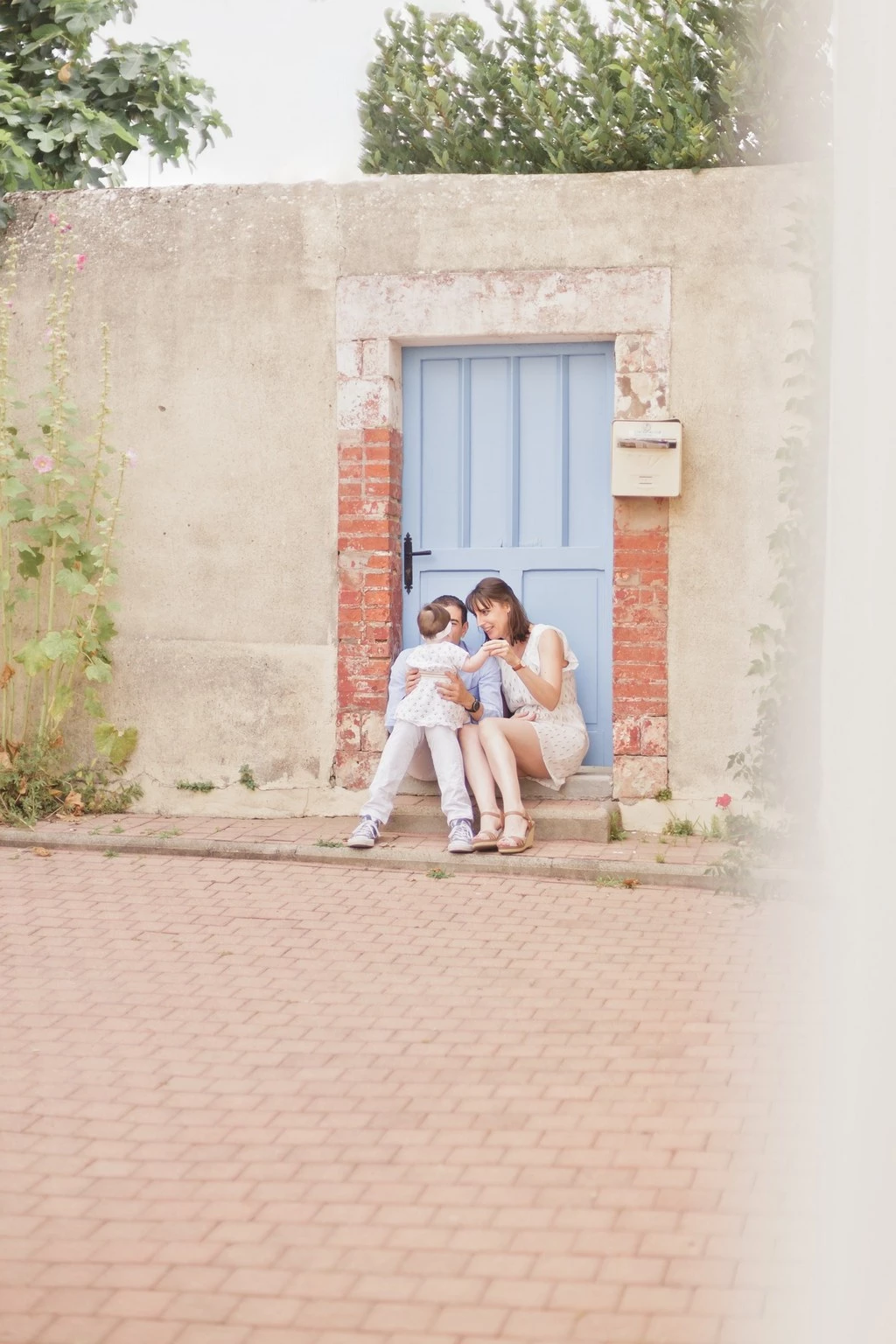 Séance photo famille : deux enfants en tenue blanche assis devant une porte bleue dans une ruelle pavée, complicité tendre et authentique