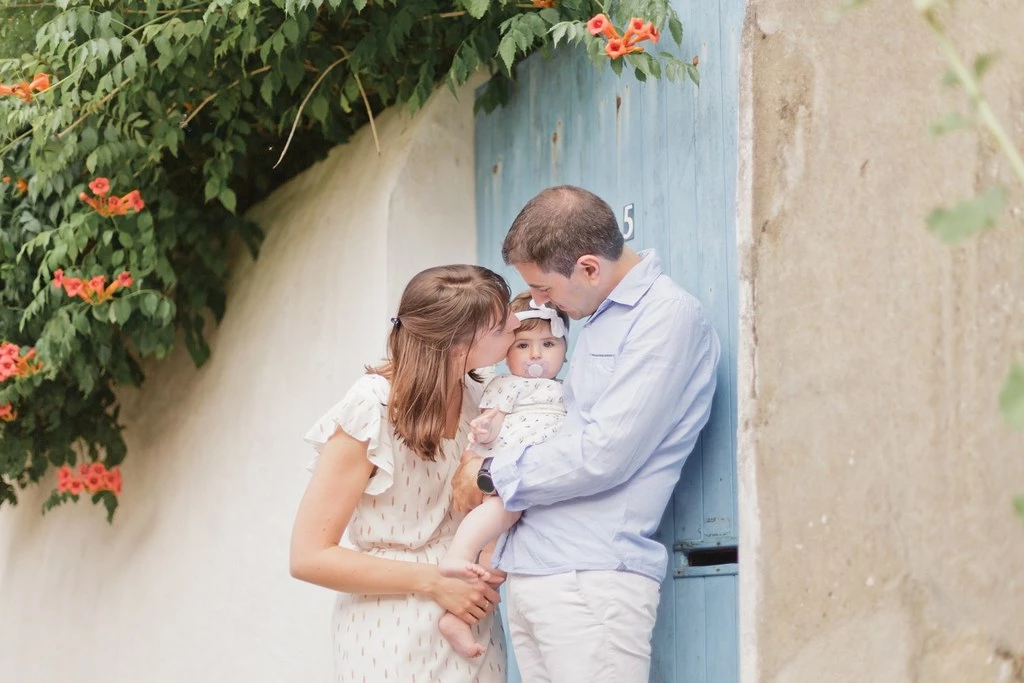 Séance photo famille tendresse : jeune couple avec bébé souriant devant porte bleue, mur blanc et lierre fleuri, lumière naturelle douce, moment d'intimité émotionnel en Vendée