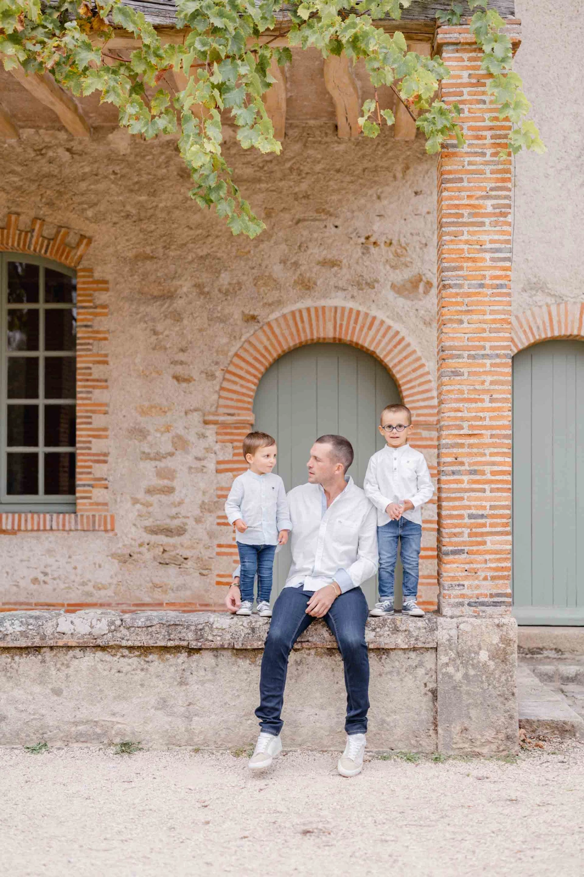Séance photo famille en extérieur : père avec deux jeunes enfants assis devant une façade en pierre et brique, ambiance tendre et complice, lumière douce dorée