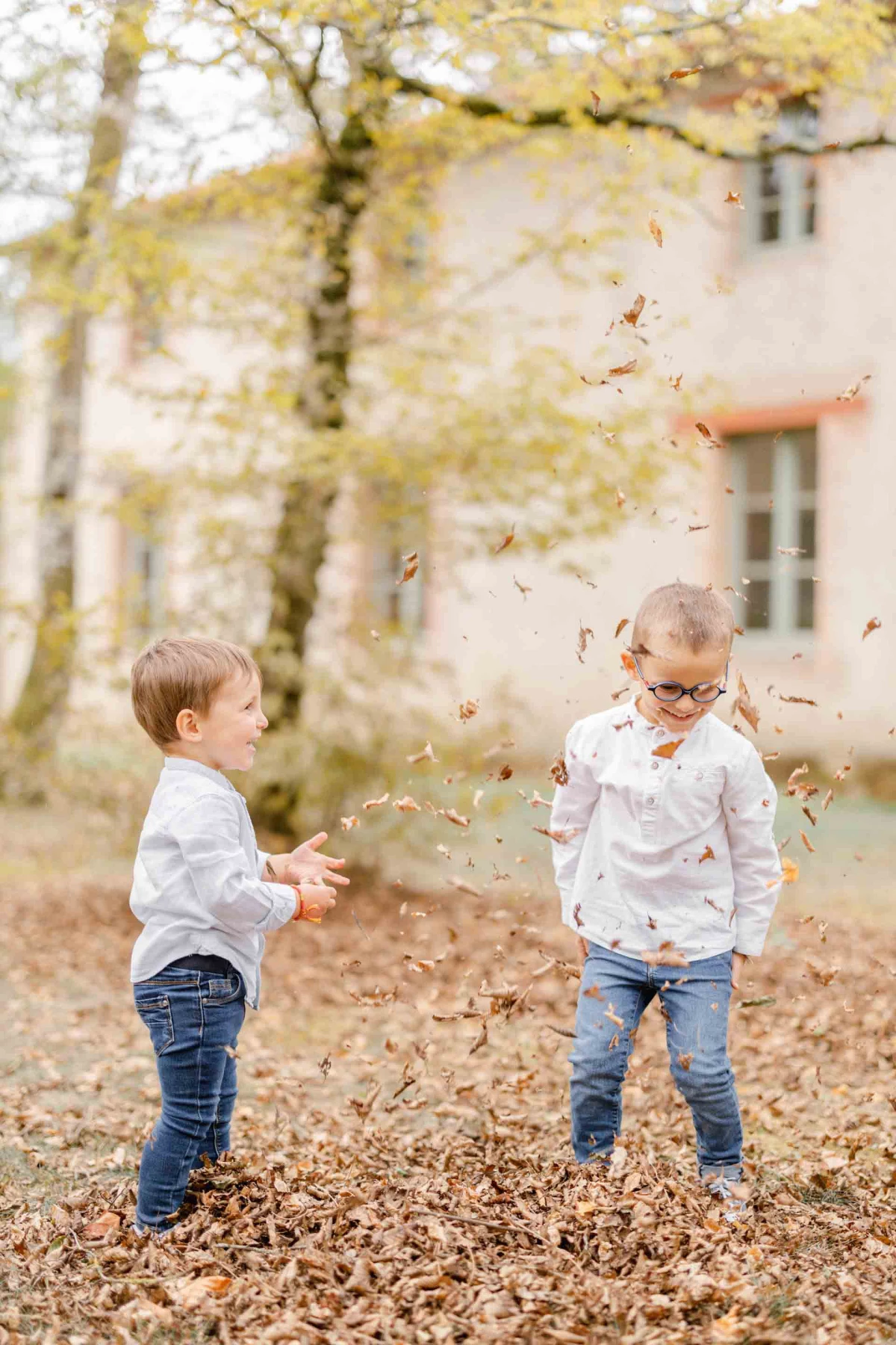 Séance photo famille automne : deux enfants jouant avec des feuilles dorées dans une cour, complicité et tendresse sous la lumière chaleureuse