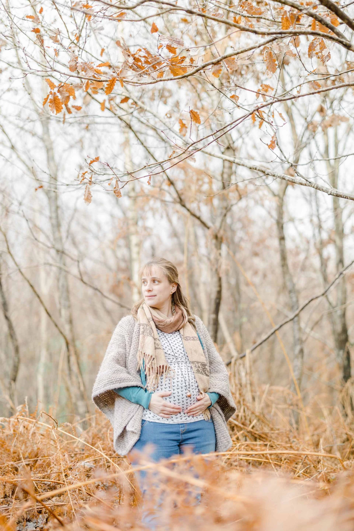 Séance photo de maternité en forêt d'automne, femme enceinte en pull beige et jean bleu, entourée de branches dorées, ambiance douce et intemporelle