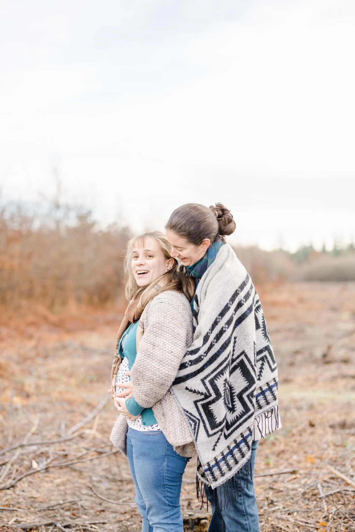Séance couple en extérieur automnal : jeune femme souriante enveloppée dans une couverture à motifs géométriques, enlacée tendrement par son compagnon, lumière douce, champs nus et arbres en arrière-plan.