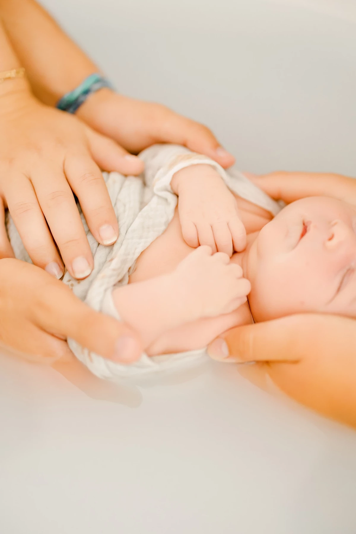 Séance bain bébé au studio : mains maternelles enveloppent un nouveau-né dans un lange blanc, instant de tendresse et de douceur.