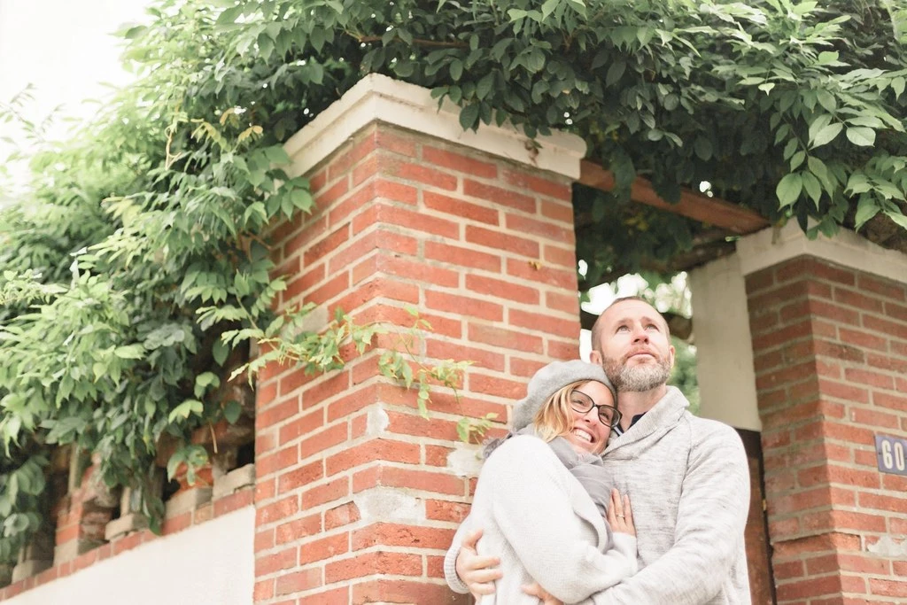 Séance famille tendresse : jeune couple enlacé devant mur brique avec lierre, lumière dorée, moment d'intimité authentique en extérieur