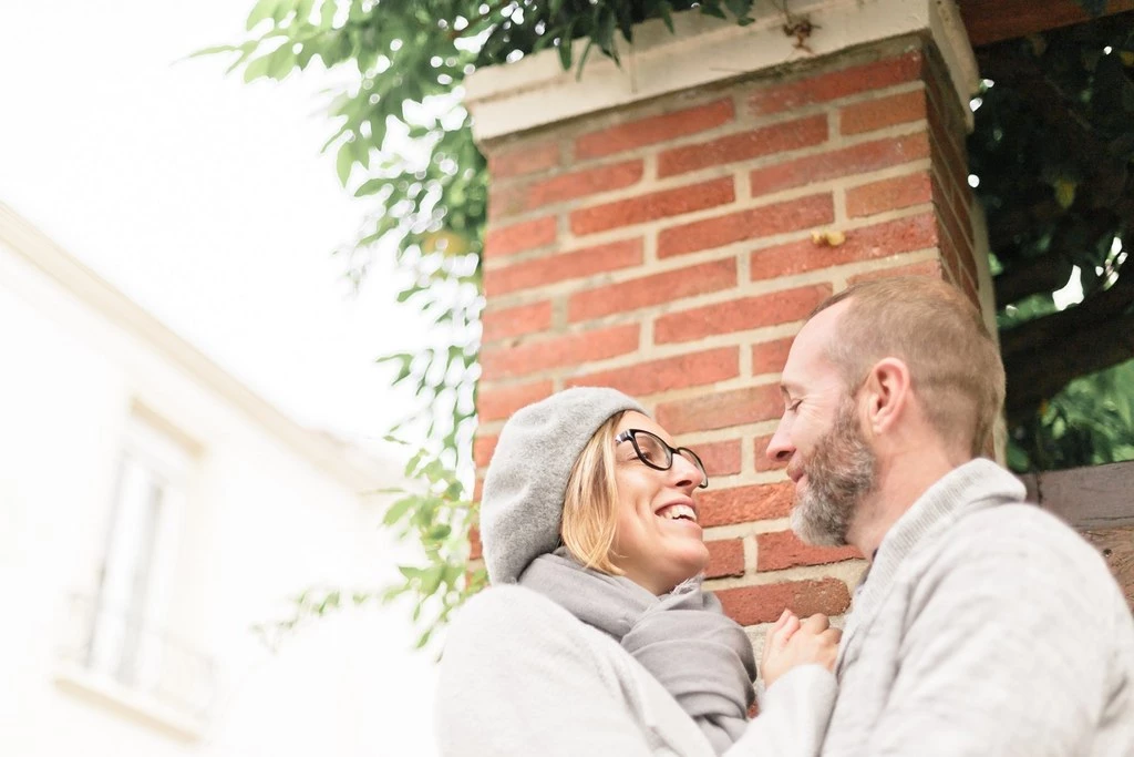 Séance couple tendresse : deux personnes enlacées devant un mur de briques rouges, échange de regards complice et sourire, lumière naturelle douce, ambiance intime et authentique