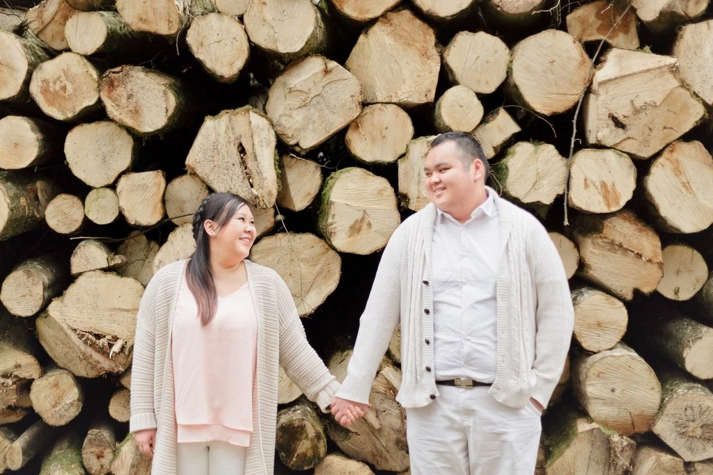 Couple complice se tenant la main devant un mur de bûches de bois, vêtements beige et blanc