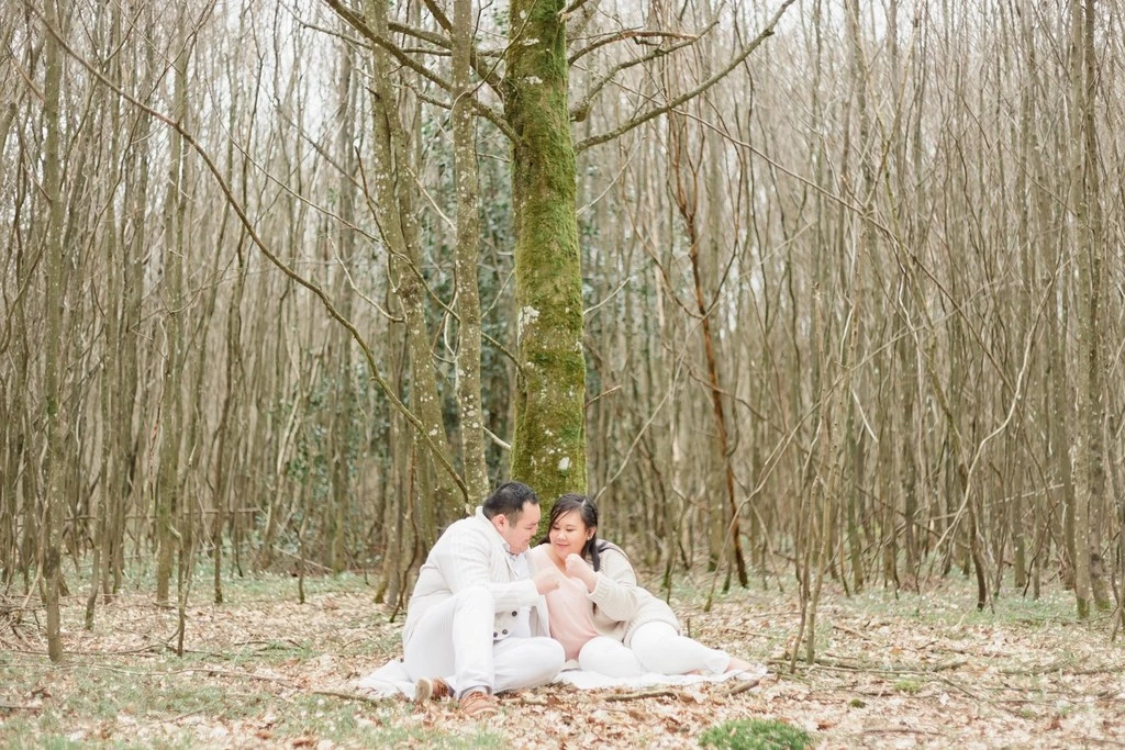Séance famille en forêt : jeunes parents et enfant assis sur un plaid blanc sous un grand arbre, ambiance tendre et intemporelle en clairière boisée