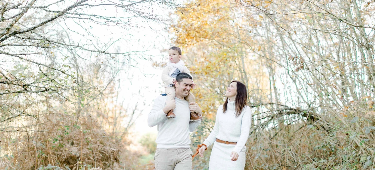Séance photo famille en automne : père portant son enfant sur les épaules, mère à ses côtés, dans une allée boisée baignée de lumière dorée, ambiance tendre et complice
