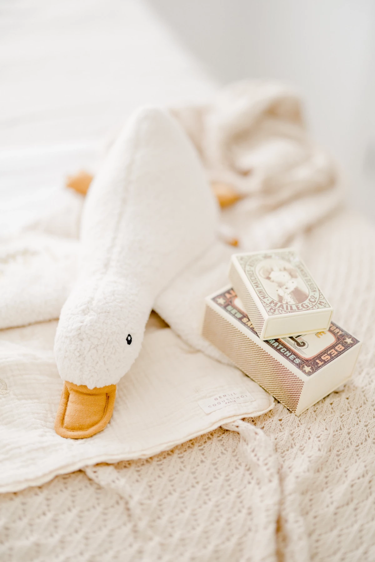 Photo d'ambiance bébé : doudou canard blanc posé sur une serviette beige avec des boites, détails tendres en studio