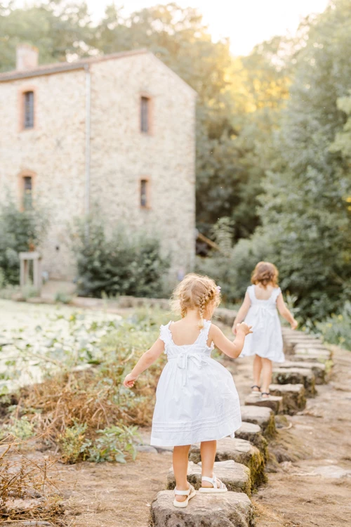 Séance photo famille : deux petites filles en robes blanches se tenant la main sur des marches en pierre, devant une maison en pierre avec jardin, lumière dorée du coucher de soleil