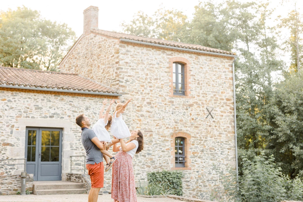 Séance famille en Vendée : père soulevant tendrement sa fille devant une maison en pierre, moment de complicité et de joie dans une lumière dorée