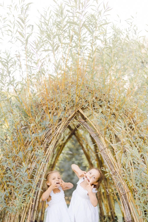 Séance photo famille avec deux petites filles en robes blanches sous une arche de roseaux dorés, moment de tendresse et complicité dans la lumière douce