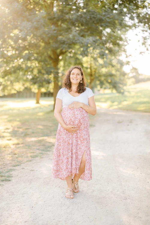 Séance de maternité en extérieur : femme enceinte vêtue d'une jupe rose fleurie caressant son ventre, sous une voûte d'arbres dorés par la lumière naturelle, dans une ambiance tendre et intemporelle