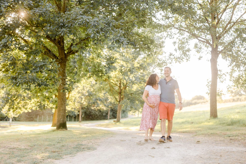 Séance famille en extérieur : couple marchant main dans la main sous une allée d'arbres centenaires, vêtus élégamment, baignés de lumière dorée et de tendresse