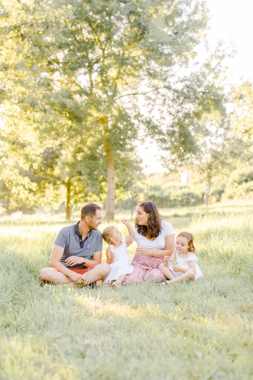 Séance photo famille en plein air : parents et deux enfants assis dans l'herbe sous la lumière dorée des arbres, moment de tendresse et complicité