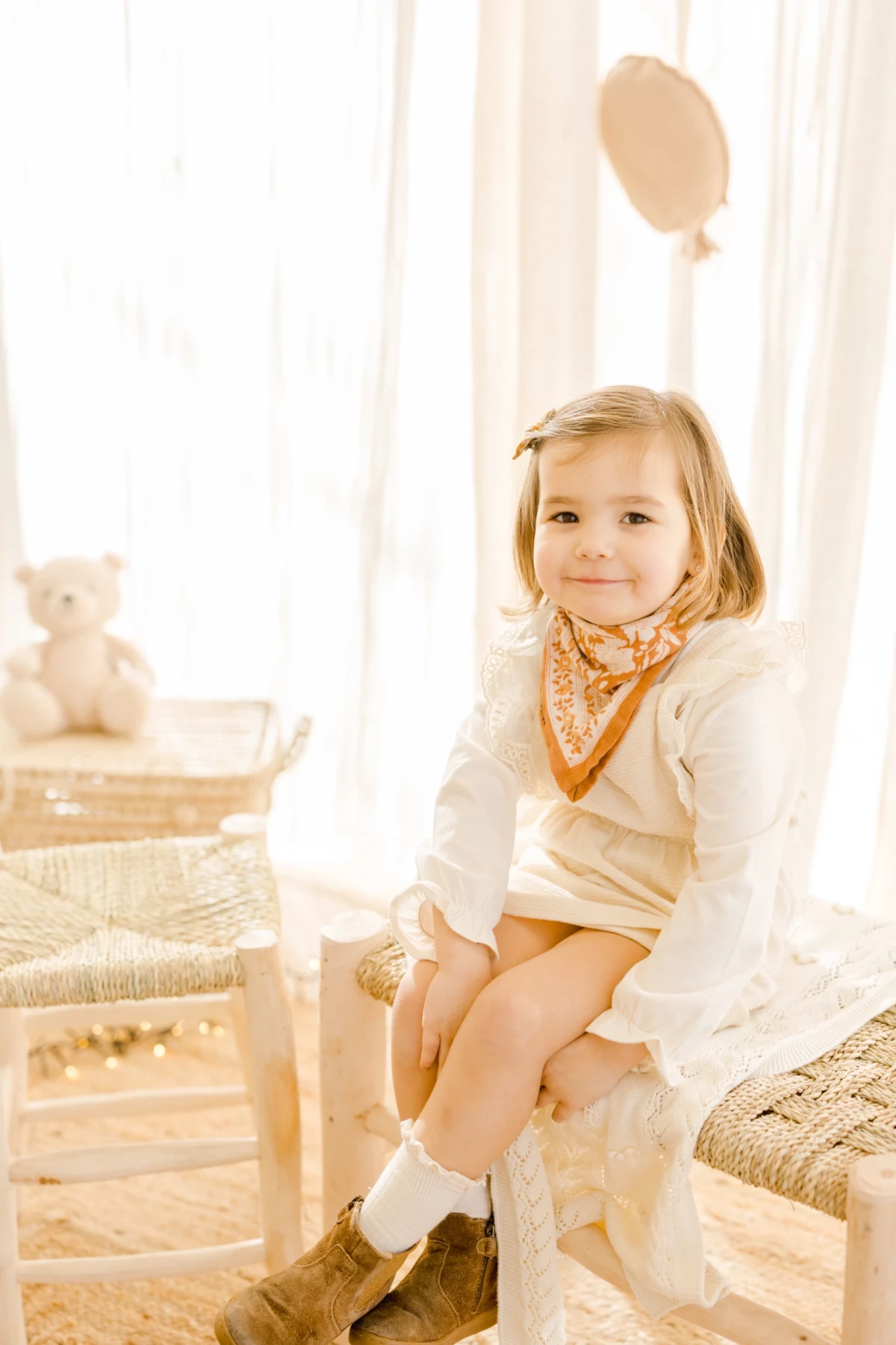 Séance enfant en studio : petite fille souriante assise sur une chaise en osier, ambiance douce et lumineuse aux tons beige