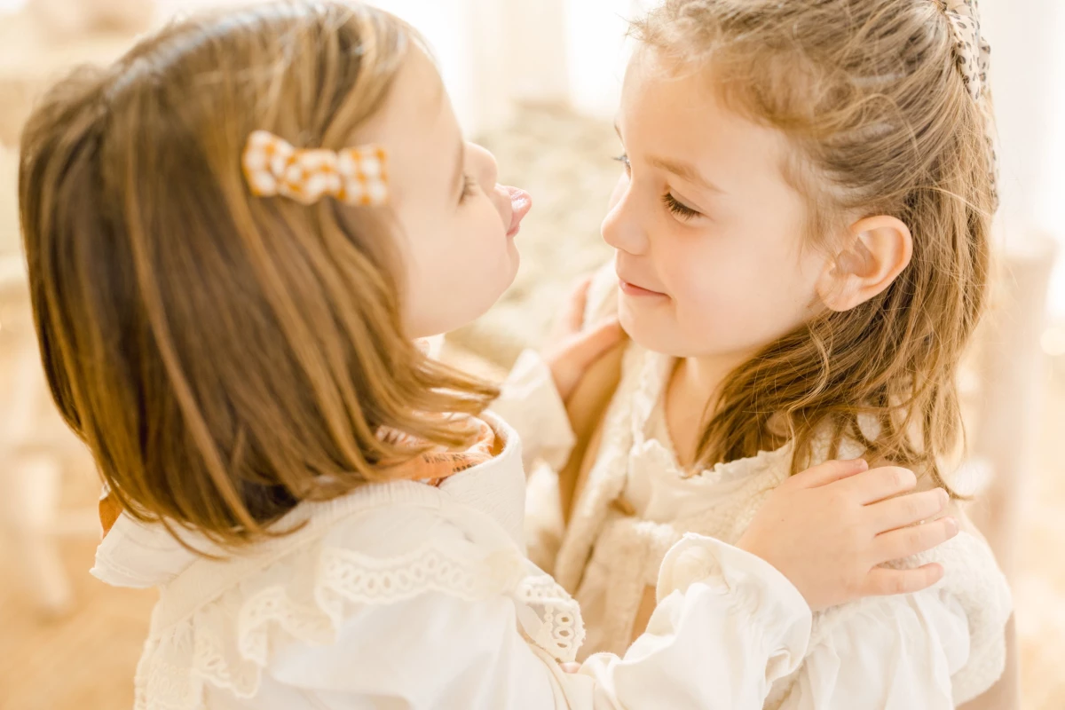 Séance famille : deux jeunes sœurs en robes blanches partagent un moment de complicité et de tendresse dans une lumière dorée