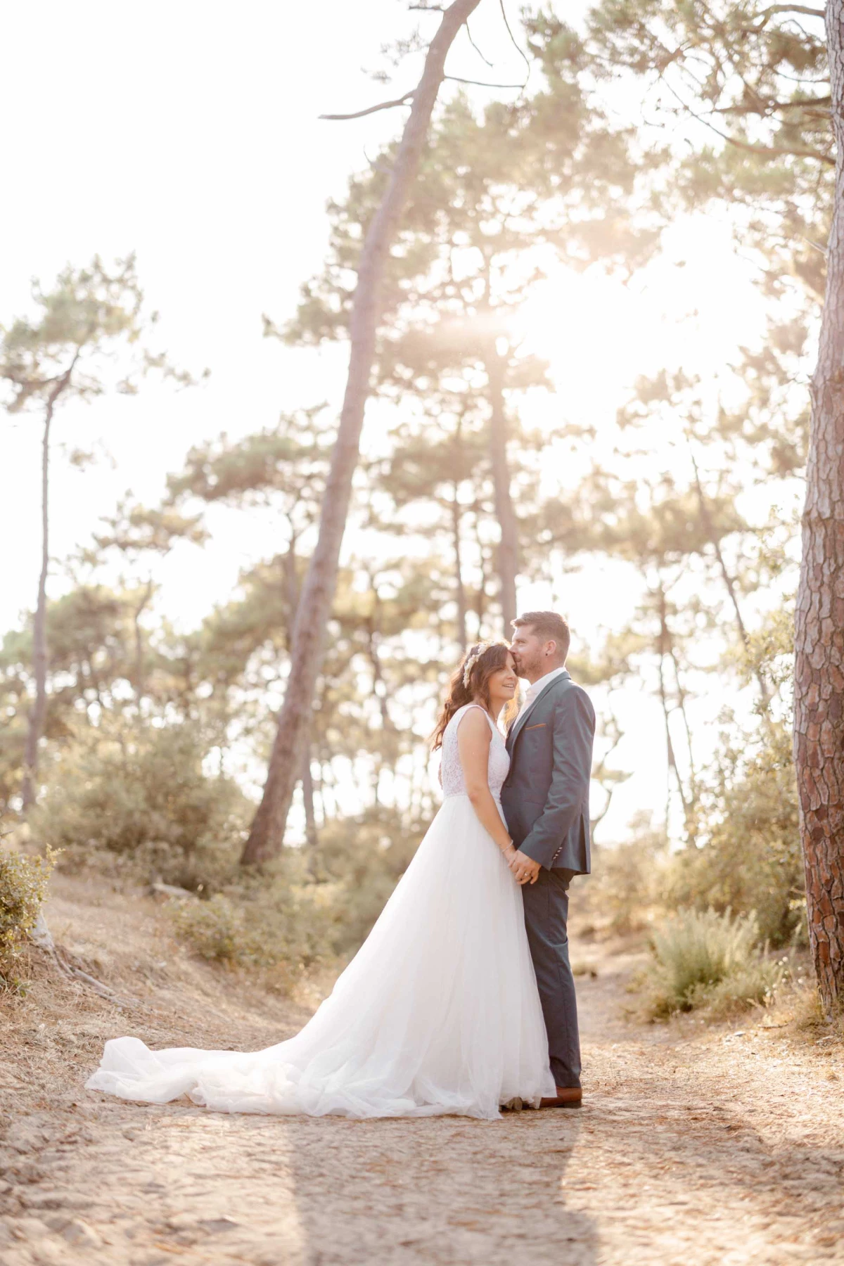 Séance photo mariage en forêt : mariée en robe blanche et marié en costume gris se regardent tendrement sous les pins dorés par la lumière du coucher de soleil