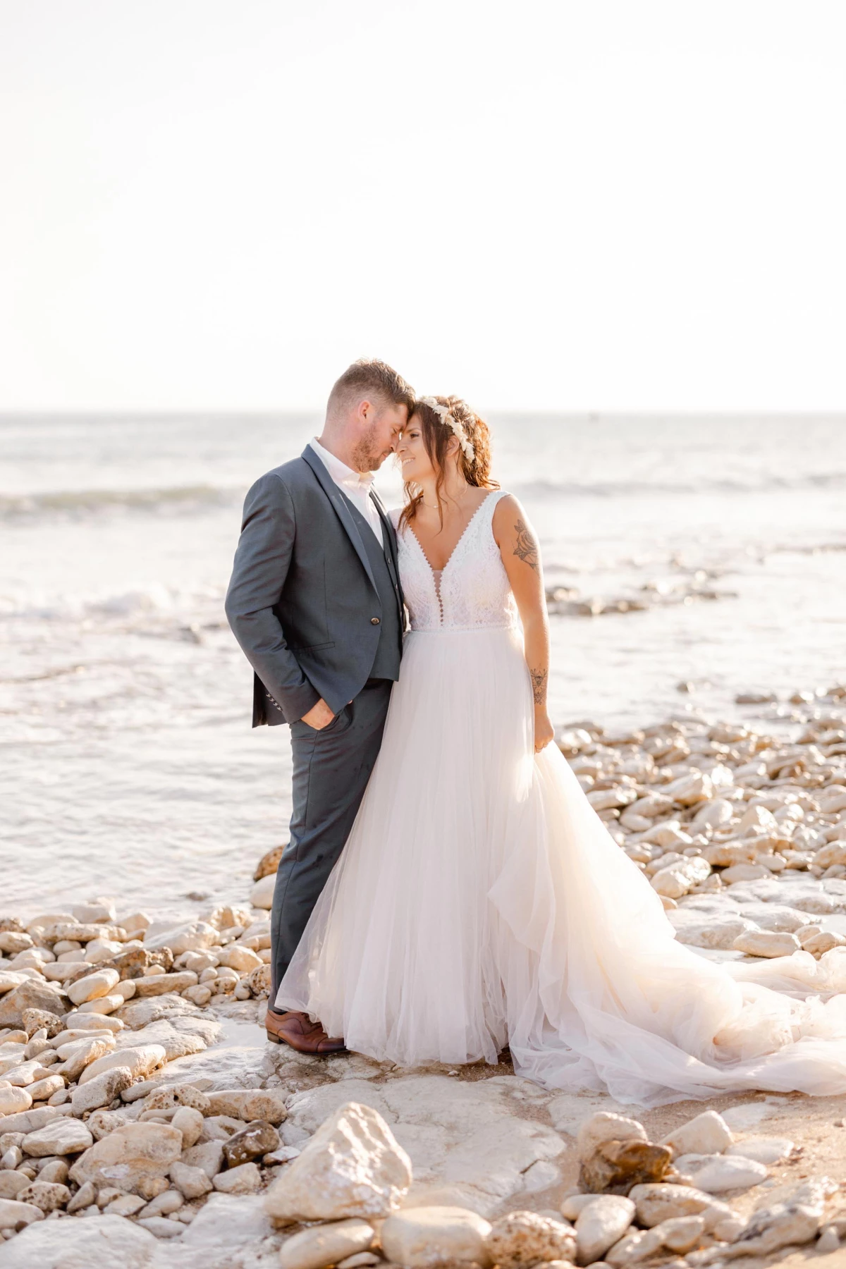Mariage à la plage : mariés s'embrassant tendrement sur galets, robe blanche fluide, costume gris, vagues en arrière-plan, lumière dorée du jour