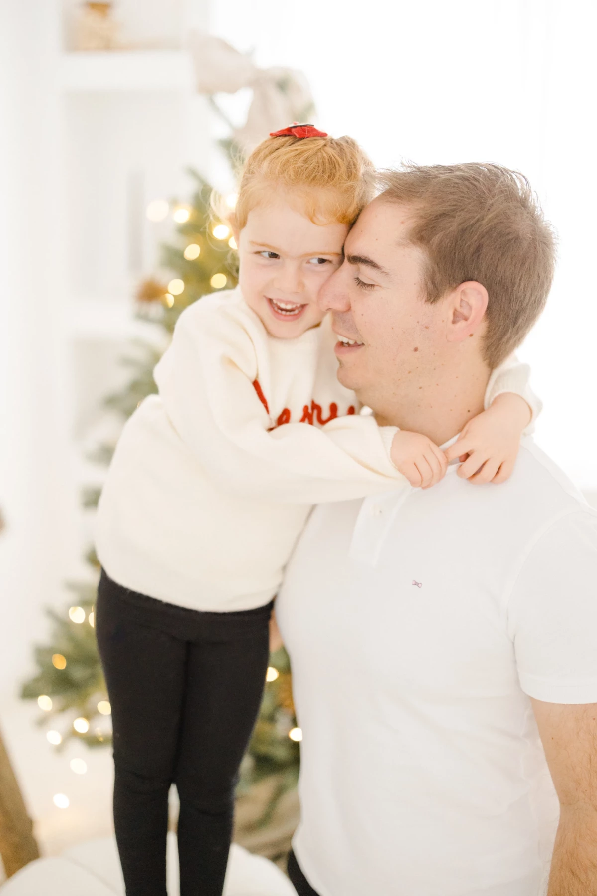 Père et fils complices devant le sapin de Noël, partageant un moment de tendresse en tenue blanche