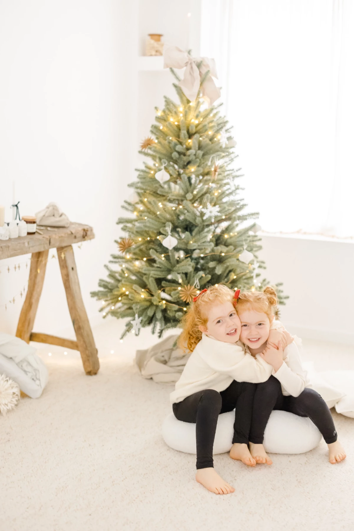 Deux enfants enlacés devant un sapin de Noël enneigé dans un studio lumineux aux tons blancs