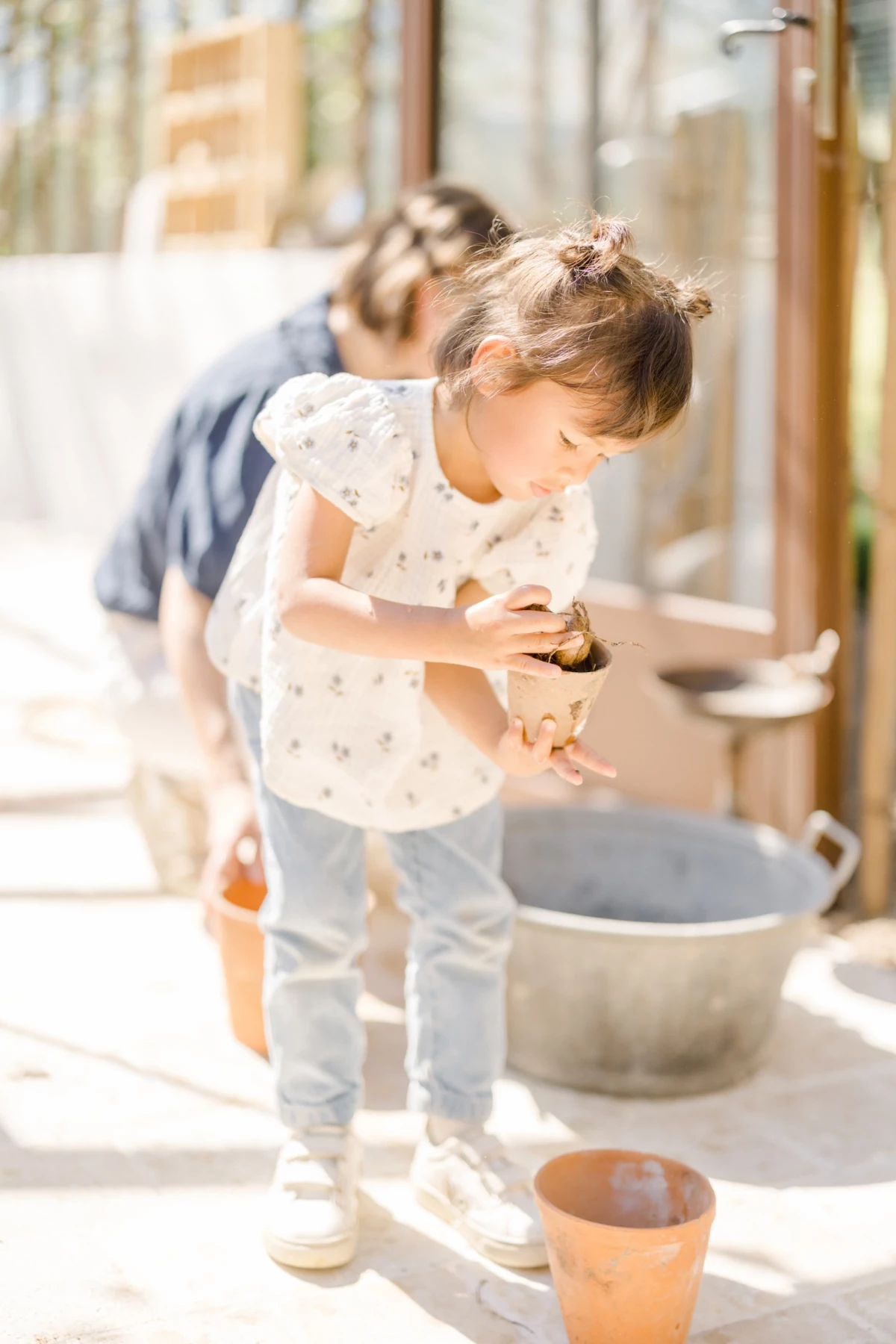 Séance famille Vendée : fillette concentrée explore la terre lors d'un atelier jardinage mère-fille dans une verrière lumineuse