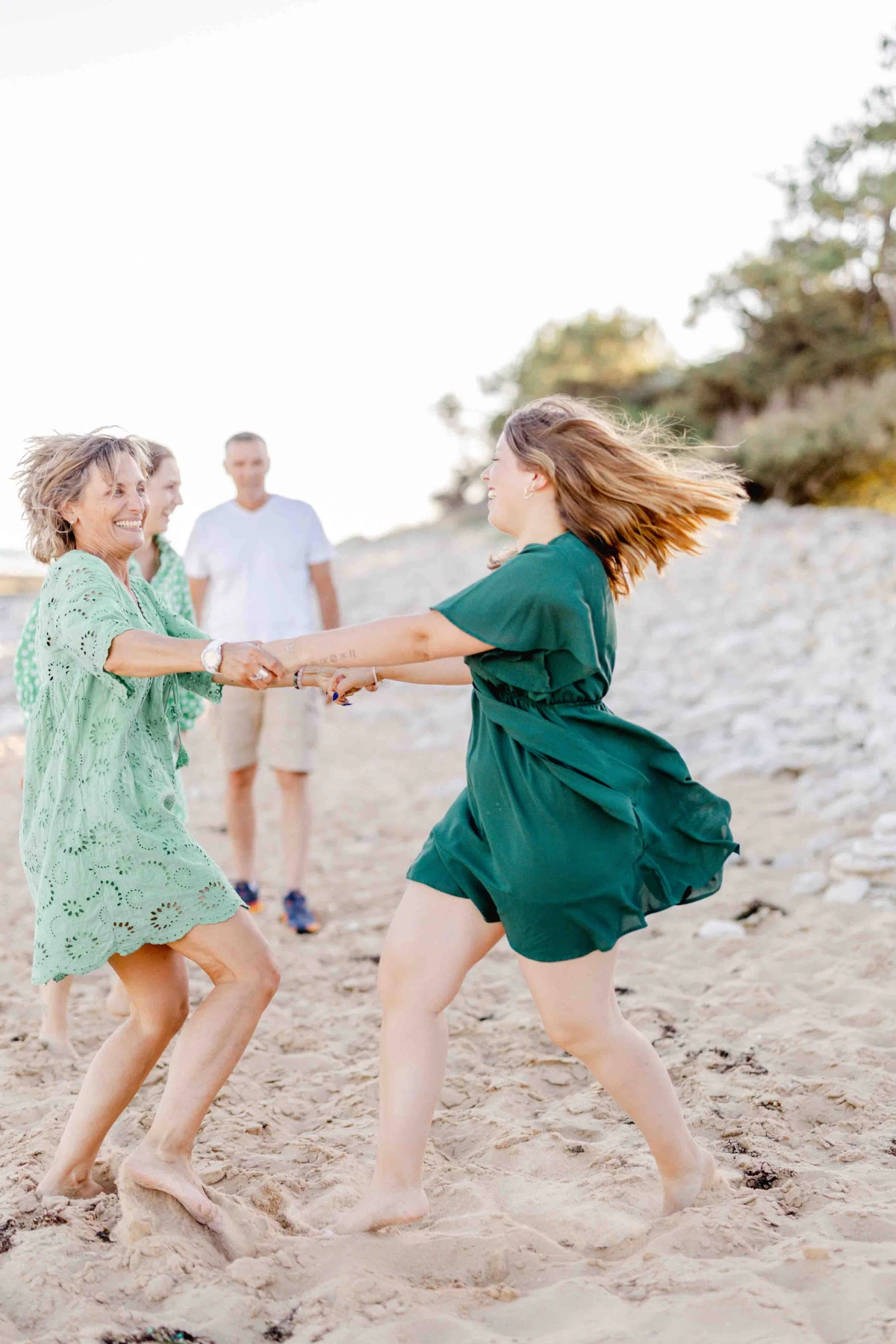 Séance famille à la plage en Vendée : deux enfants se tenant les mains dans une danse joyeuse sur le sable, avec un parent en arrière-plan, lumière dorée et ambiance tendre