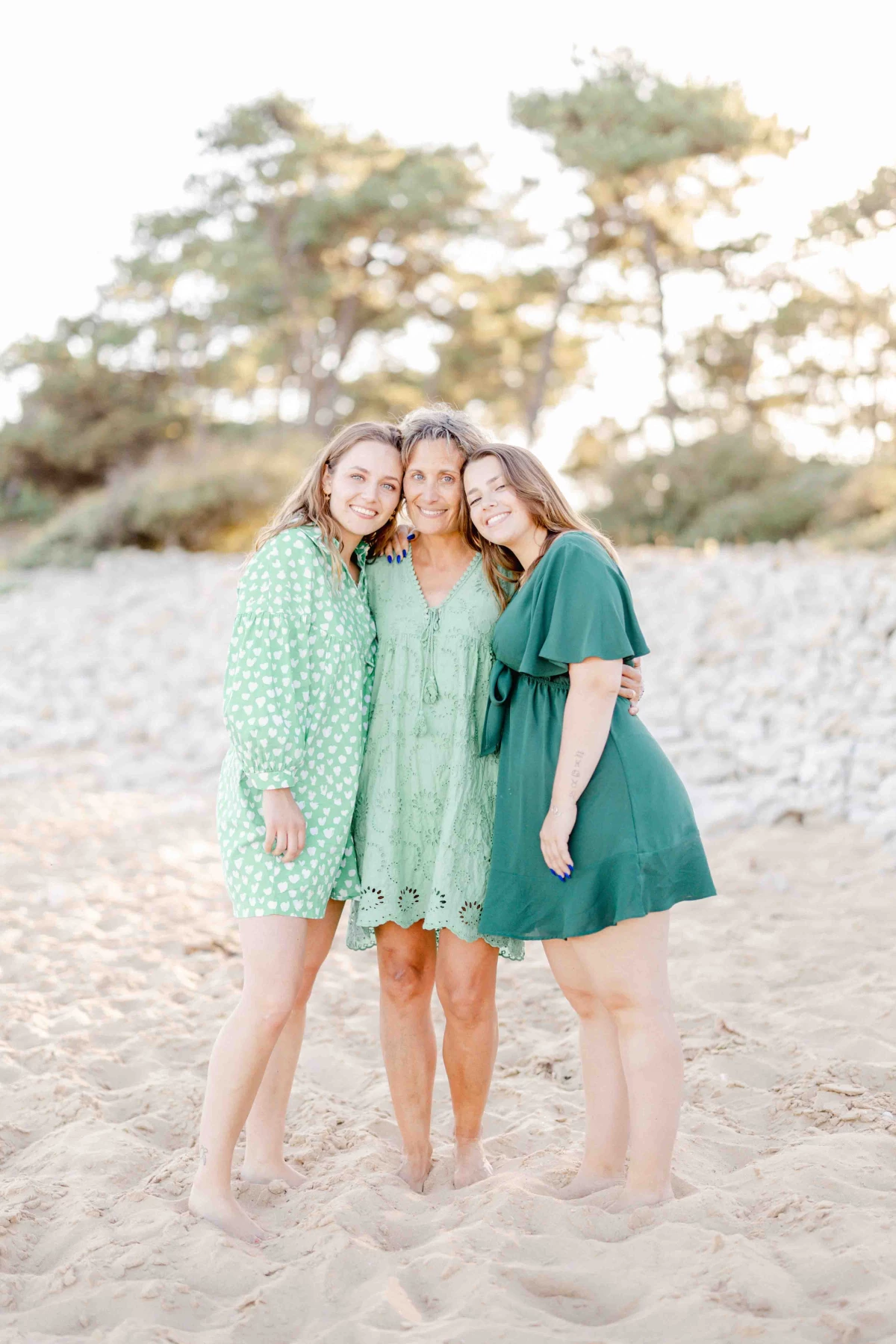 Séance photo famille sur la plage : trois femmes en robes vertes posent avec tendresse et complicité face à la caméra, baignées de lumière dorée, avec pins parasols en arrière-plan