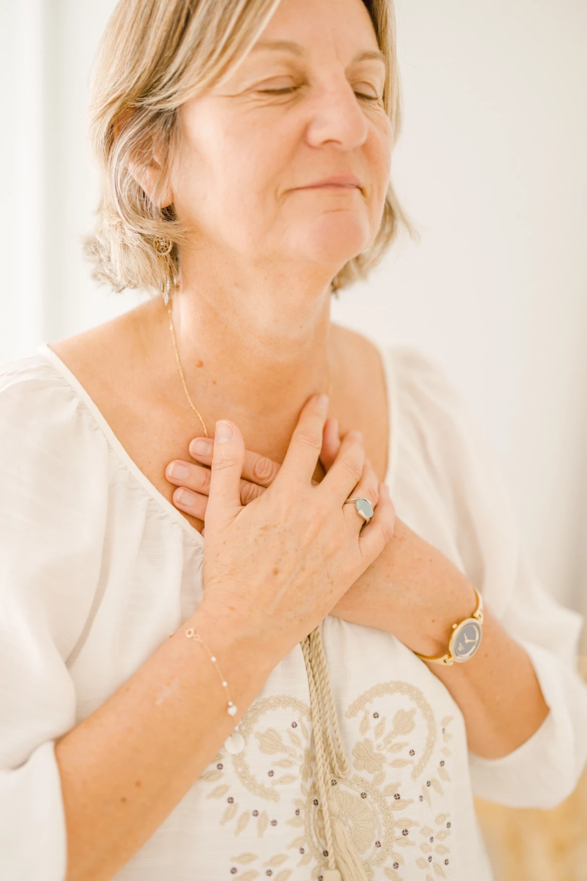 Femme blonde en chemisier blanc, les mains sur le cœur, les yeux fermés dans un geste de gratitude