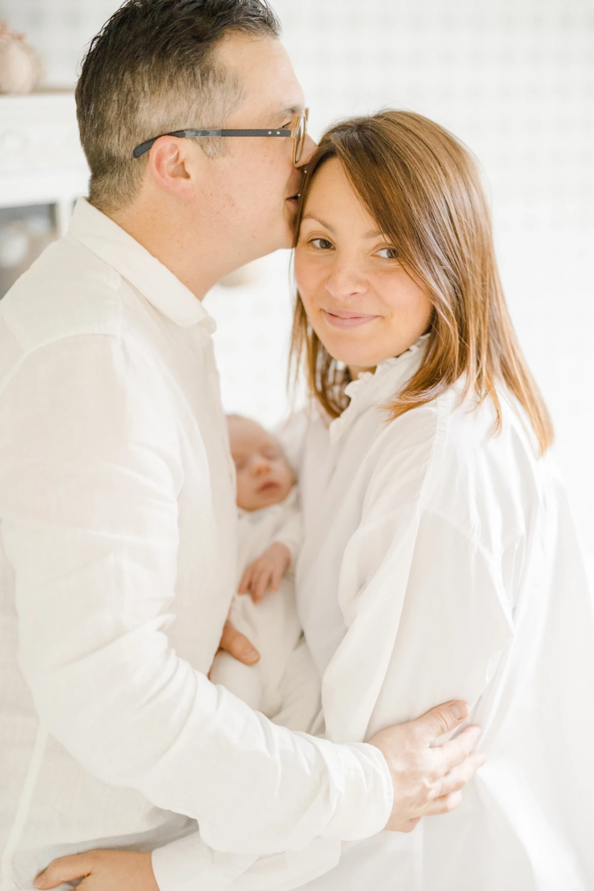 Séance famille en studio : parents enlacent leur enfant dans une ambiance de douceur et complicité, tout de blanc vêtus