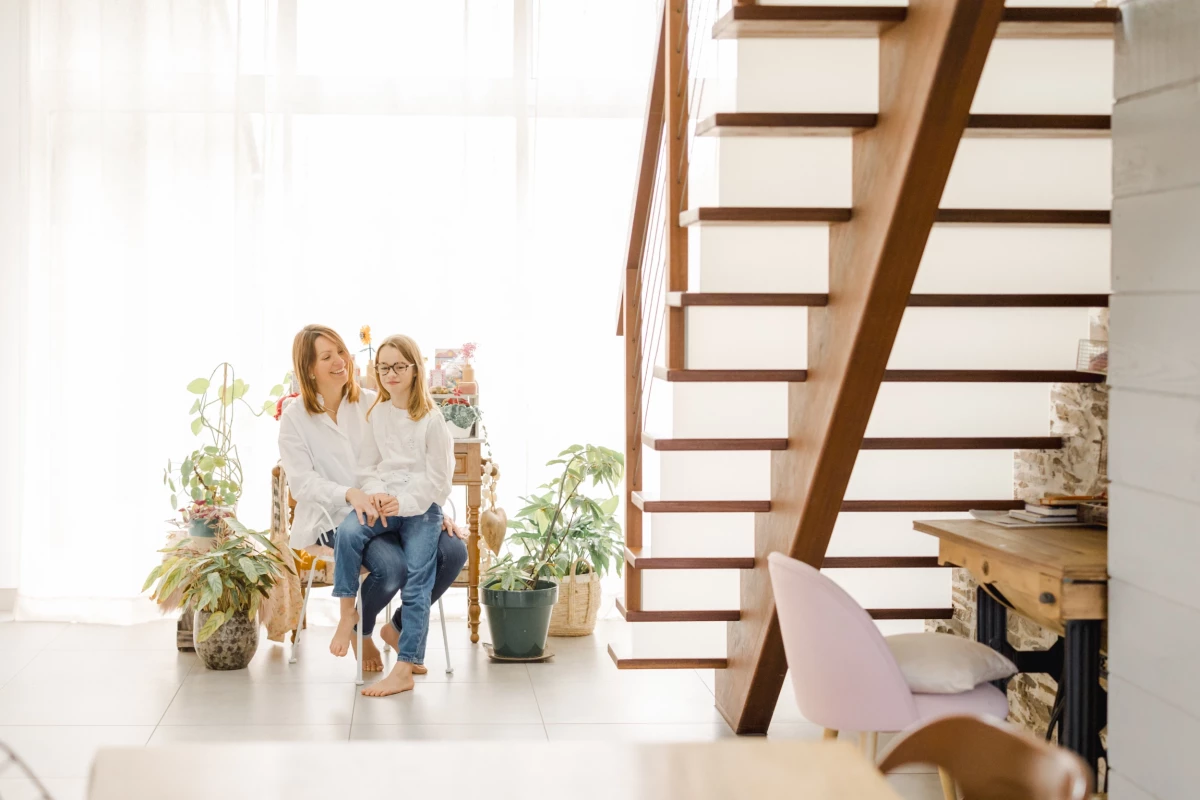 Séance famille au domicile : maman et ses deux filles partagent un moment de complicité au pied d'un escalier en bois, lumière douce