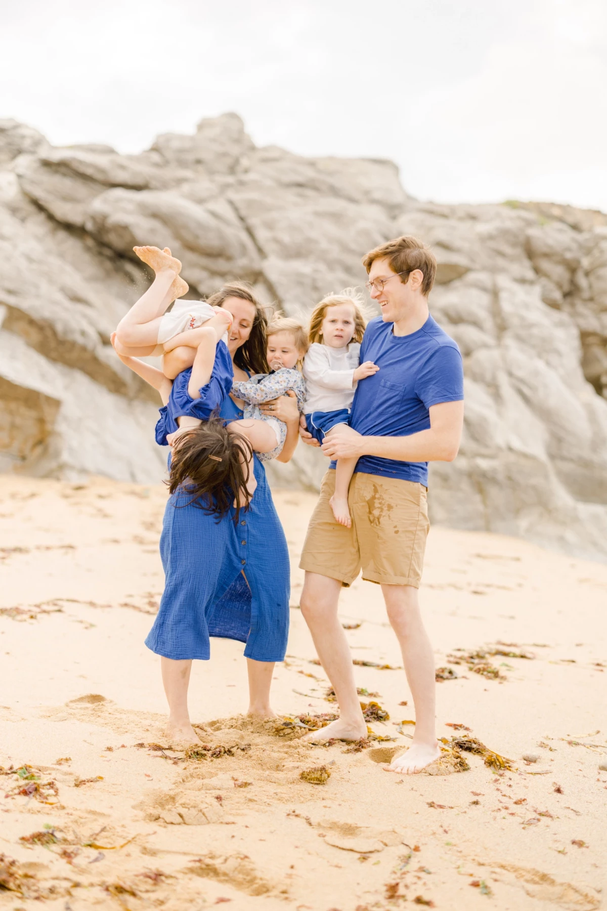 Famille de quatre personnes en tenues bleues assorties posant sur une plage de sable, rochers en arrière-plan