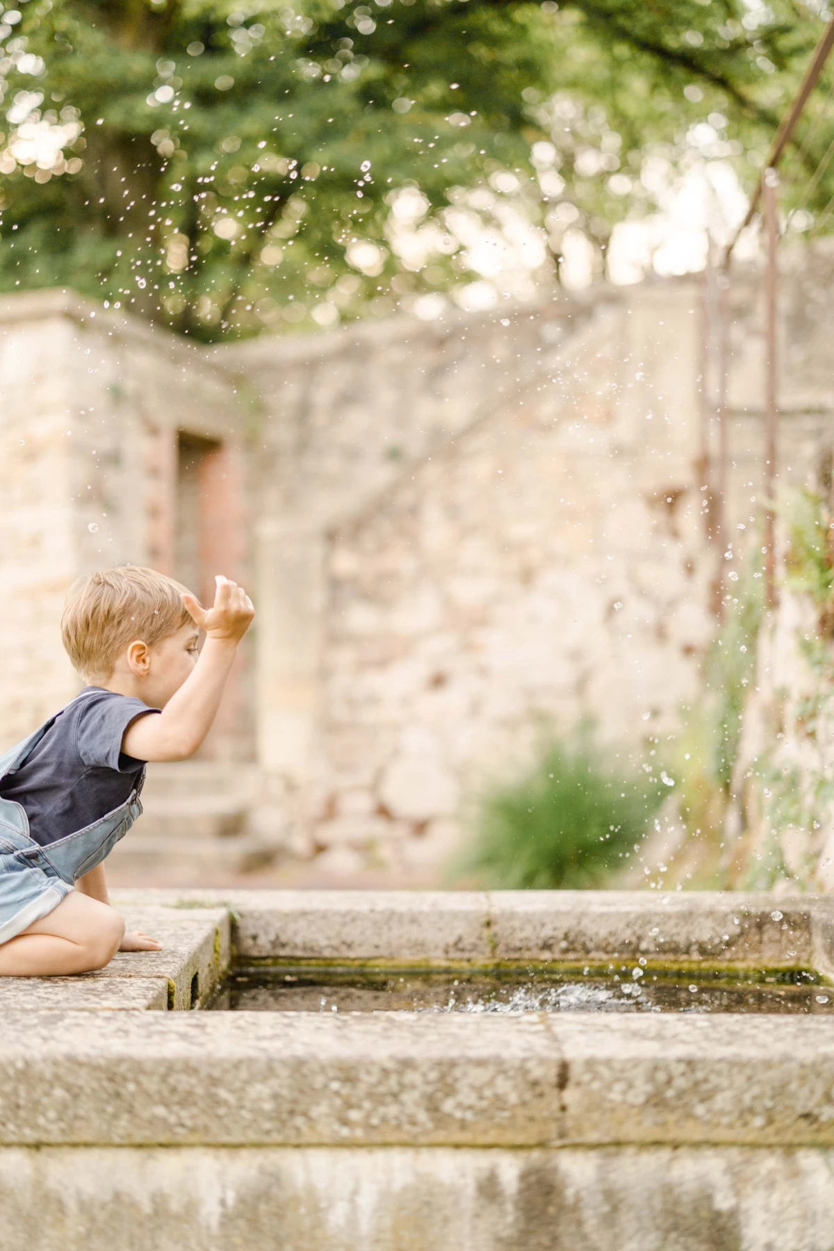 Petit garçon blond jouant avec l'eau d'une fontaine en pierre dans un jardin de château