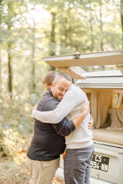 Séance famille en van aménagé : père et fils s'étreignent tendrement devant un camping-car rétro dans une clairière forestière ensoleillée, moment d'authenticité et complicité