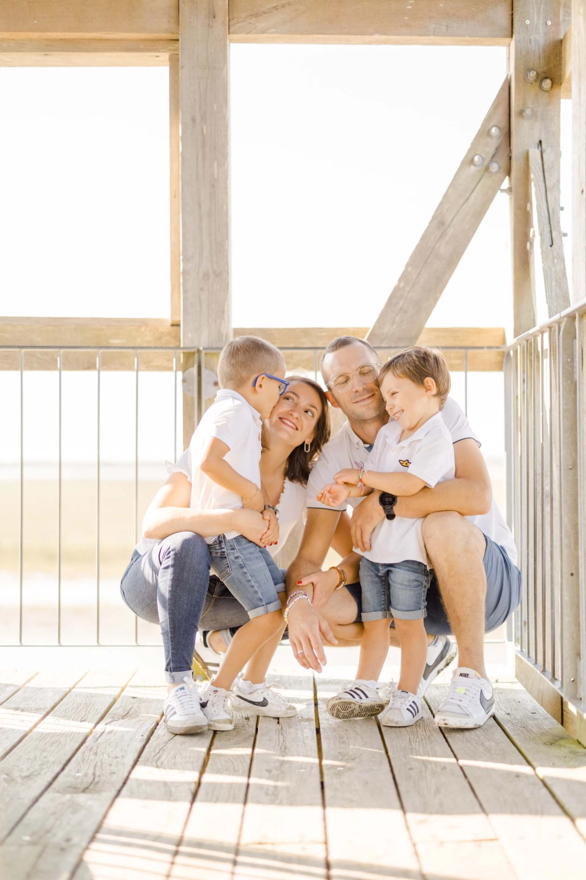 Famille de quatre personnes enlacée sur une terrasse en bois, vêtus de blanc, baignés de lumière dorée