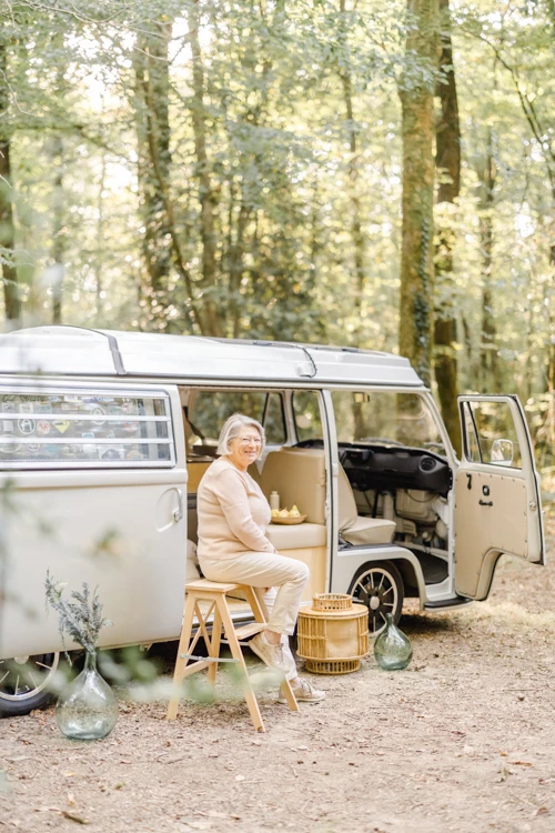 Portrait de femme senior en camping-car vintage dans une forêt ensoleillée, moment de sérénité et d'authenticité en pleine nature