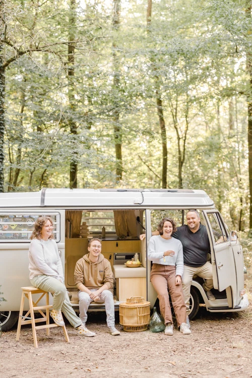 Séance famille lifestyle en van aménagé en forêt, quatre adultes souriants partageant un moment de complicité au cœur de la nature, lumière dorée filtrée par les arbres.