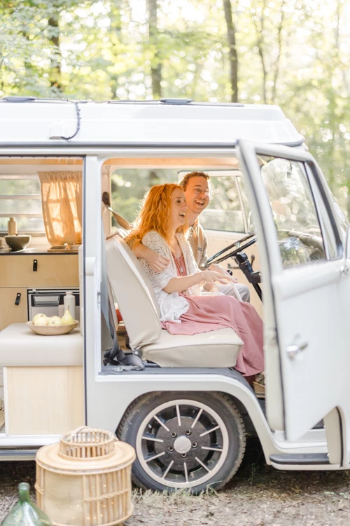 Séance famille dans un camping-car vintage blanc : une mère et sa fille rient ensemble au volant, lumière dorée filtrant par les vitres, ambiance intemporelle et chaleureuse en forêt