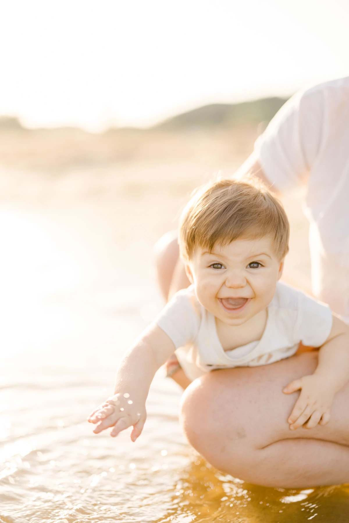 Bébé souriant tenu au-dessus de l'eau sur une plage au coucher du soleil