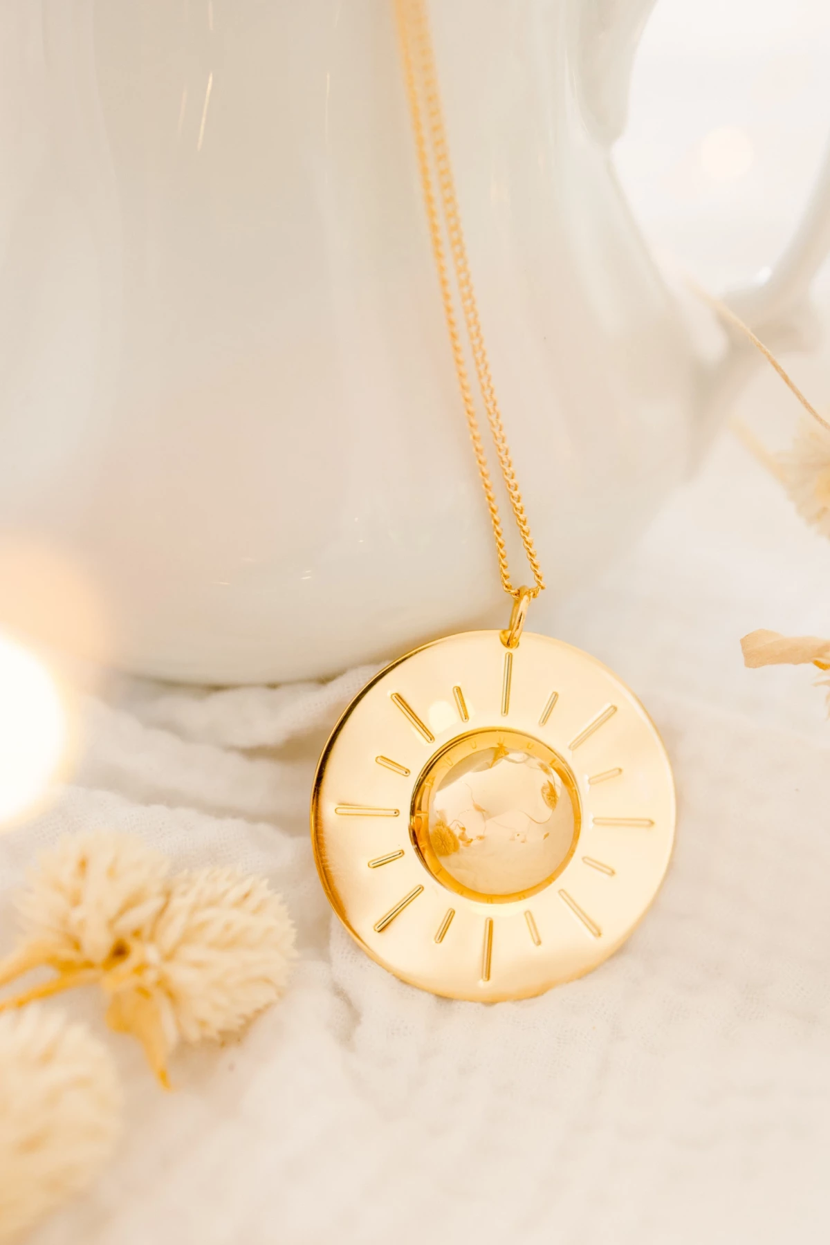 Collier doré avec pendentif soleil posé sur surface blanche entouré de fleurs séchées beiges