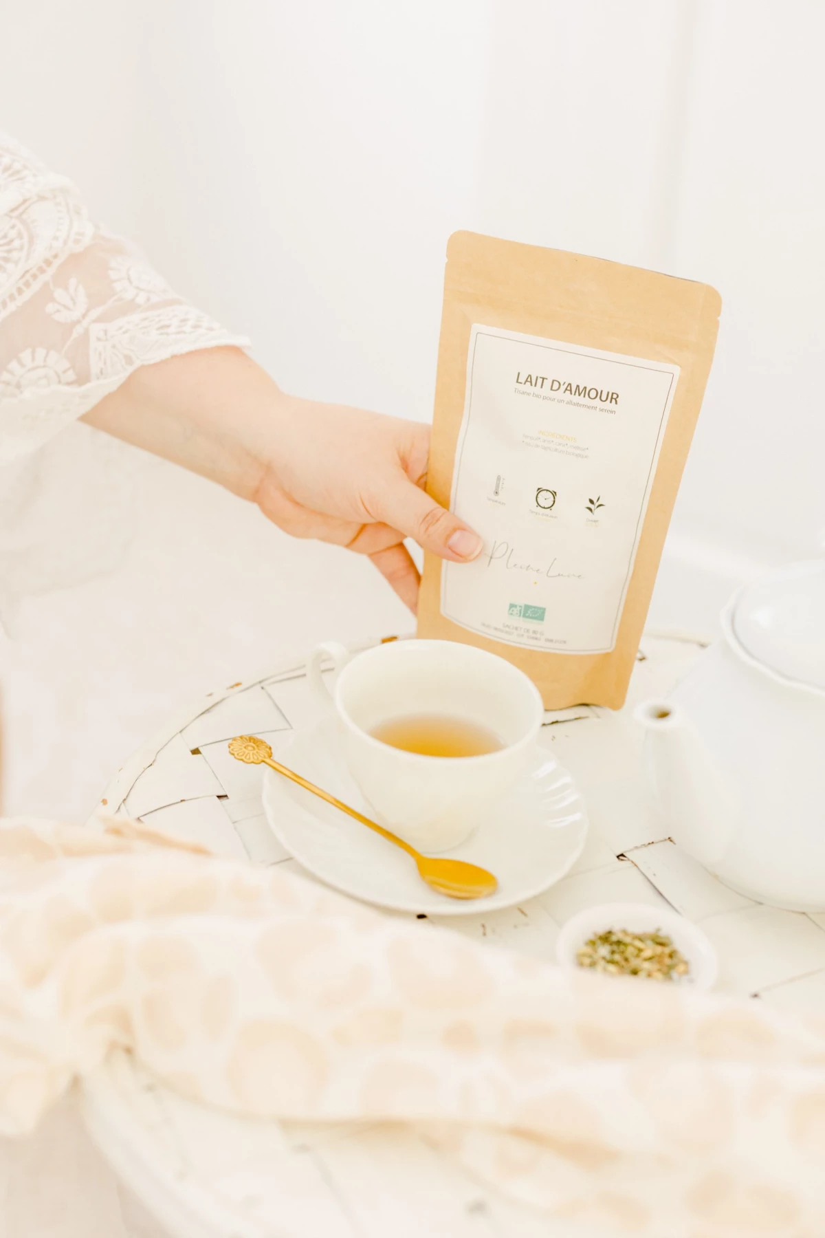 Main féminine tenant un sachet de tisane Lait d'Amour près d'une tasse de thé et d'une cuillère dorée