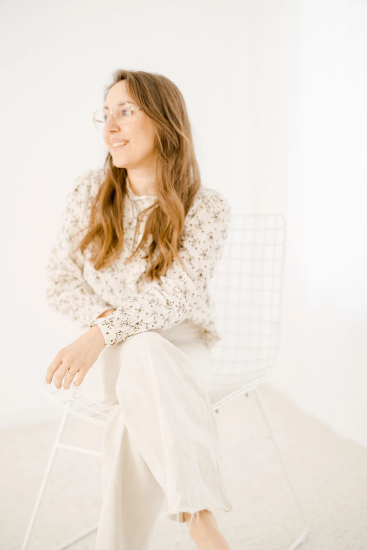 Portrait lumineux d'une femme souriante en robe blanche fleurie, assise sur une chaise dans un studio épuré