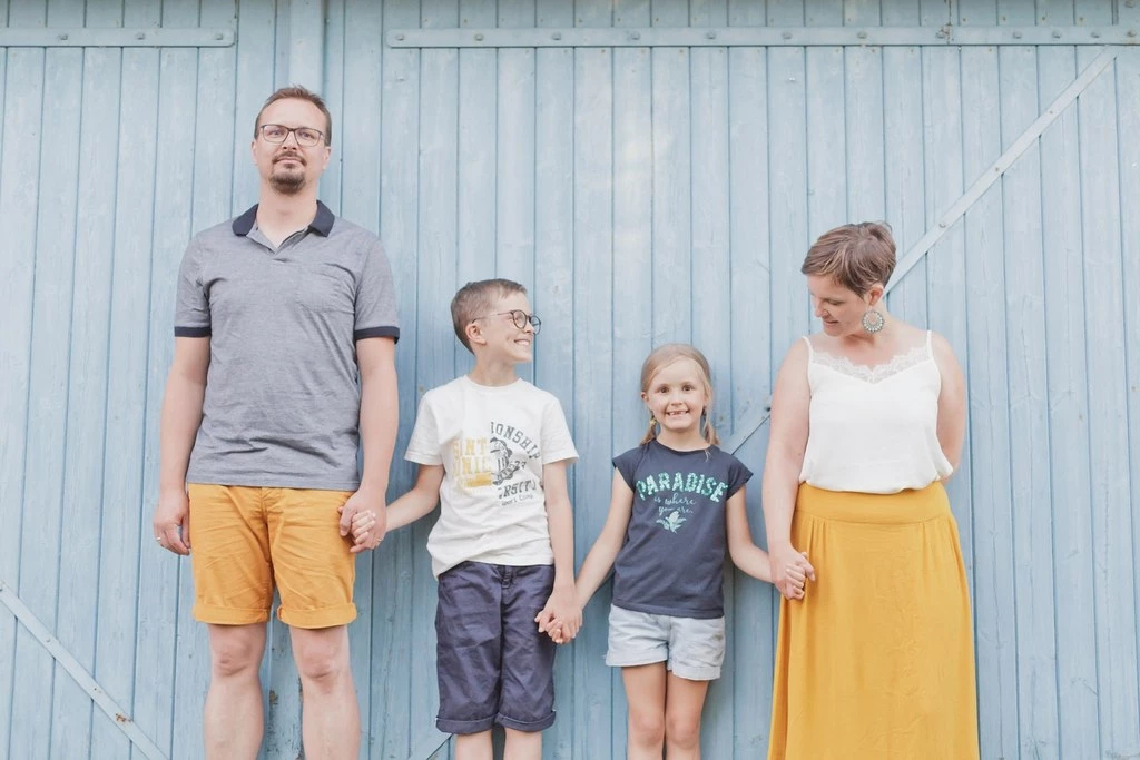 Séance photo famille : quatre personnes souriantes se tenant la main devant un mur bleu ciel, ambiance douce et complice, lumière naturelle et intemporelle