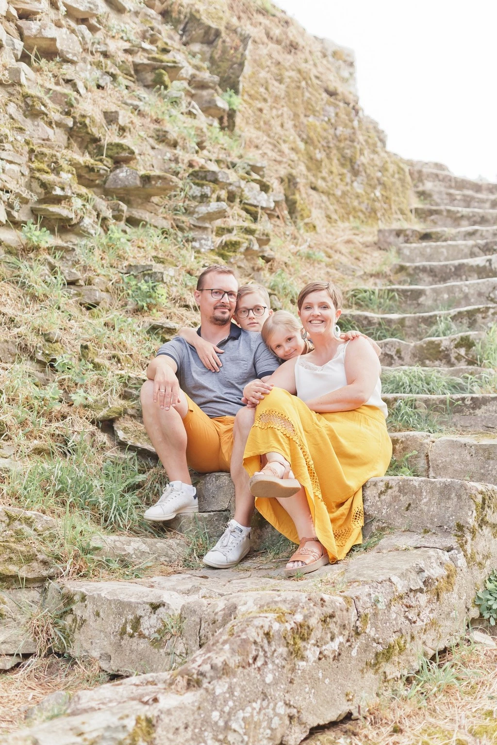 Séance photo famille en extérieur : parents et deux enfants en tendre complicité sur des escaliers en pierre, vêtus de couleurs chaudes, ambiance douce et intimiste