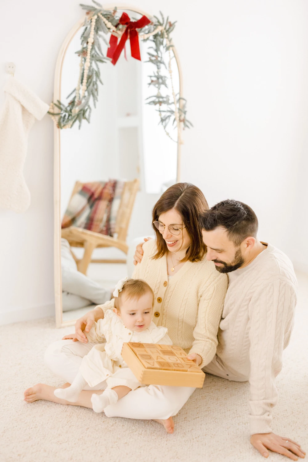 Séance photo famille intemporelle : parents et bébé en tenues neutres partagent un moment de tendresse sous une arche décorée, lumière douce et naturelle, ambiance chaleureuse et intime.